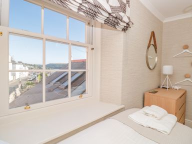 A bedroom window with a view over rooftops and a street next to a wooden nightstand with a lamp and folded towels at Upper Tremorvah Dartmouth