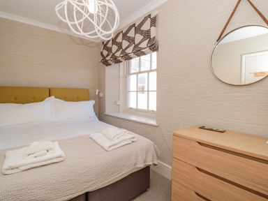 A bedroom with a bed with white linens and folded towels by the window with patterned curtains and a wooden dresser with a round mirror above at Upper Tremorvah in Dartmouth