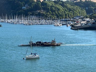 A harbor with a small sailboat in the water and a ferry carrying vehicles near a marina with many docked sailboats and a tree-covered hill in the background at Upper Tremorvah in Dartmouth