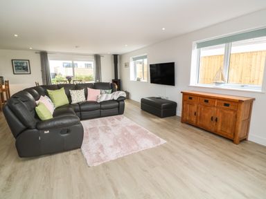 A living room with a black sectional sofa with cushions a wooden sideboard a wall mounted tv a small wood burning stove and large windows at 3 Praa Cove in Praa Sands