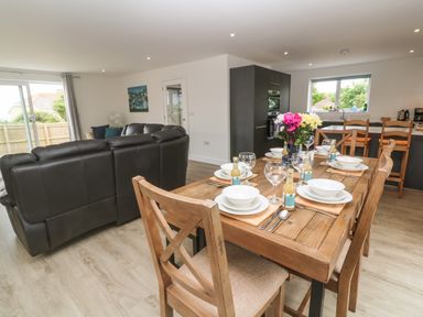A dining area with wooden table set with plates glasses and bottles next to a living room with leather sofas and a kitchen with a window at 3 Praa Cove in Praa Sands
