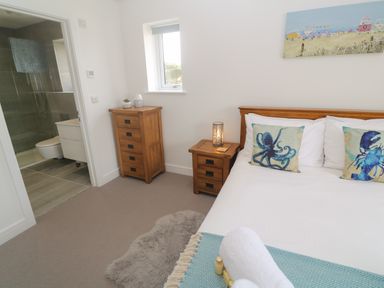 A bedroom with a wooden bed with ocean-themed cushions a wooden bedside table with a lamp a wooden chest of drawers a window and a bathroom entrance at 3 Praa Cove in Praa Sands