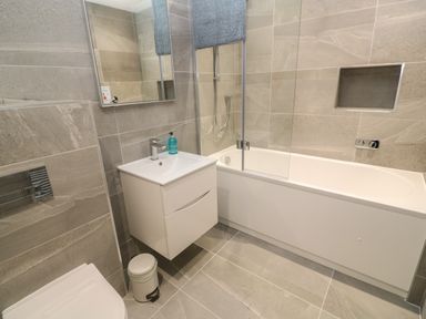 A bathroom with a white bathtub with glass screen a white sink with cabinet a mirror a toilet and a trash bin at 3 Praa Cove in Praa Sands