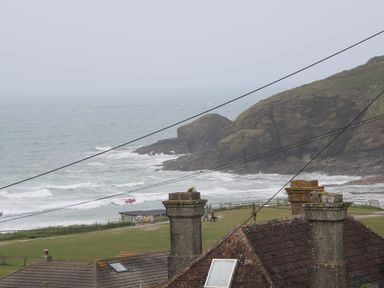 A coastal scene with rooftops and chimneys in the foreground a grassy area with people walking and a flag near the shore and rocky cliffs by the sea at 3 Praa Cove in Praa Sands