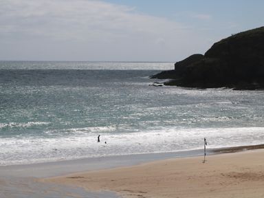 A beach with two people in the water and a flag on the sand near rocky cliffs at 3 Praa Cove in Praa Sands