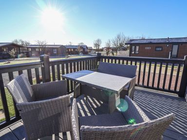 An outdoor wooden deck with a table and four wicker chairs overlooking a holiday park with multiple cabins at Owl's Nest in Newton on Derwent near Wilberfoss