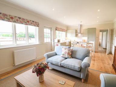 A living room with a blue sofa and a wooden coffee table with flowers and a kitchen and dining area in the background at Flagstone Cottage in Upper Swell near Stow-On-The-Wold