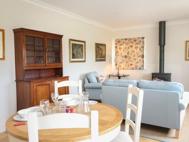 A dining table with chairs and tableware in a living room with sofas and a wood stove at Flagstone Cottage Upper Swell near Stow-On-The-Wold