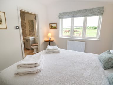 A bedroom with a white bedspread and folded towels on the bed a bedside table with a lamp and a window showing horses outside at Flagstone Cottage in Upper Swell near Stow-On-The-Wold