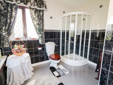 A bathroom with a shower enclosure toilet with wooden seat a small round table with flowers and patterned curtains at Derriens Farmhouse in Letterbreen County Fermanagh