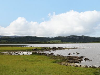 A lake with rocks and grass in front of hills under a partly cloudy sky at Derriens Farmhouse in Letterbreen County Fermanagh