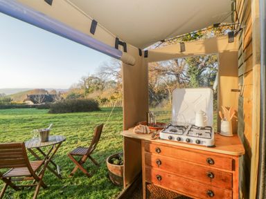 An outdoor kitchen setup with a gas stove on a wooden dresser and a small wooden table with two chairs on grass at The Wagon at Burrow Hill in Ottery St Mary