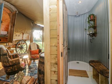 An outdoor area with wooden barrel chairs and a covered wagon beside a shower with corrugated metal walls and a wooden bench at The Wagon at Burrow Hill in Ottery St Mary