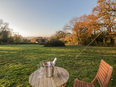 A grassy outdoor area with a wooden table and chair a bottle in an ice bucket and two glasses at The Wagon at Burrow Hill in Ottery St Mary