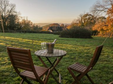 A wooden table with a metal ice bucket and two glasses on a grassy field with two wooden chairs at The Wagon at Burrow Hill in Ottery St Mary