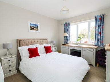 A bedroom with a bed with two red cushions bedside tables with lamps a dresser under a window with blue curtains at The Lobster Pot in Overstrand