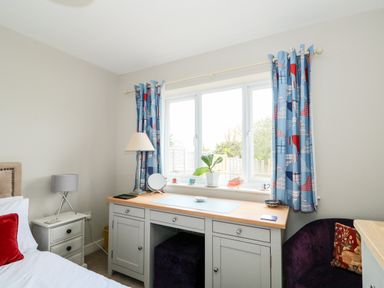 A bedroom with a bed side table lamp a desk with a mirror plant and chair by a window with blue patterned curtains at The Lobster Pot in Overstrand