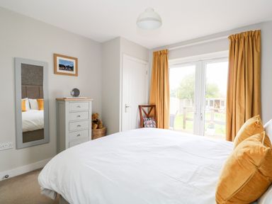 A bedroom with a white bed and yellow pillows a chest of drawers a chair and glass doors with yellow curtains at The Lobster Pot in Overstrand