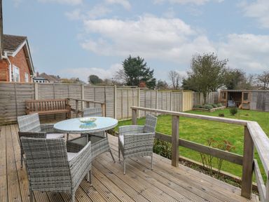 A wooden deck with a glass round table and four wicker chairs overlooking a fenced garden with grass and a garden shed at The Lobster Pot in Overstrand