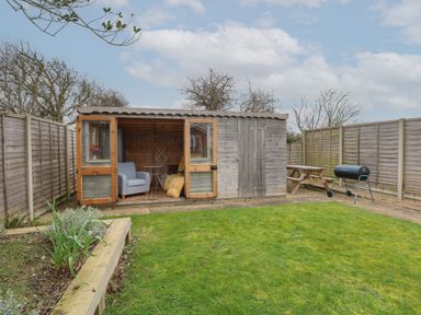 A wooden garden shed with open doors showing seating and a table a picnic bench barbecue and fence panels at The Lobster Pot in Overstrand