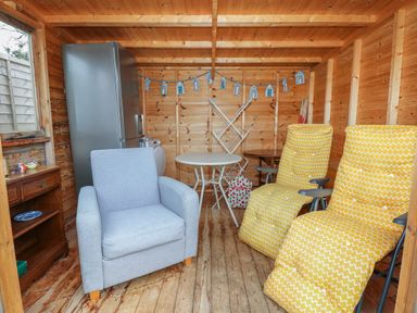 A wooden room with a gray armchair two yellow reclining chairs a round table a refrigerator and a small wooden cabinet at The Lobster Pot in Overstrand