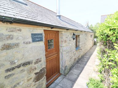 Stone exterior wall with wooden door and small window along a paved path beside green hedges at Godolphin Barn Ashton near Rosudgeon