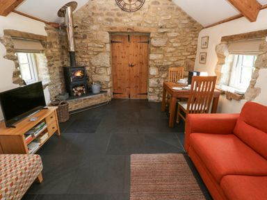 A rustic living room with a wood burning stove dining table orange sofa and television at Godolphin Barn in Ashton near Rosudgeon
