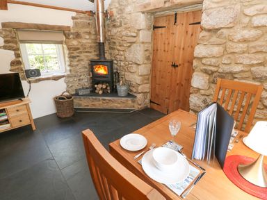 A dining area with a wooden table and chairs near a wood stove and stone walls at Godolphin Barn in Ashton near Rosudgeon