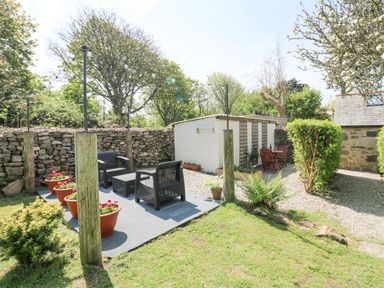 An outdoor seating area with black chairs and flower pots next to a stone wall and a small white building at Godolphin Barn Ashton near Rosudgeon