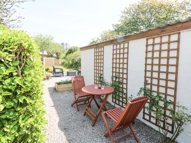 An outdoor seating area with a wooden table and two chairs next to a white wall with trellises and climbing plants at Godolphin Barn in Ashton near Rosudgeon