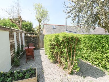 An outdoor garden area with a gravel path a wooden table and chair set green hedges and a flower bed at Godolphin Barn in Ashton near Rosudgeon