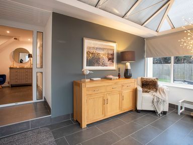 A room with a wooden sideboard, a framed picture above it, a lamp, and a white armchair with a cushion and blanket at Hafod Heli in Rhosneigr