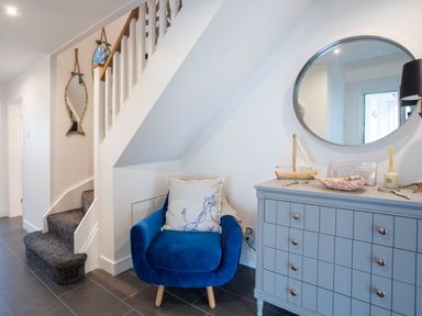 A blue armchair with cushions under stairs next to a gray dresser with a round mirror above it at Hafod Heli in Rhosneigr