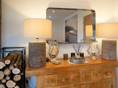 A wooden sideboard with two lamps logs in a metal rack and decorative items in a room at Hafod Heli in Rhosneigr