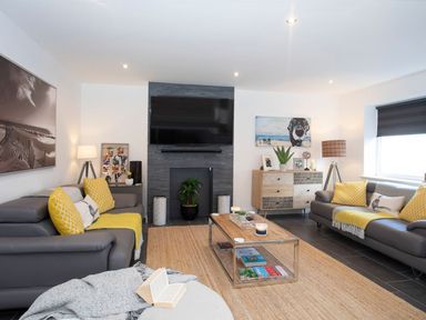 A living room with two gray sofas with yellow cushions a TV mounted above a fireplace a wooden coffee table and a sideboard at Hafod Heli in Rhosneigr