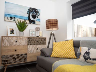 A living room corner with a patterned wooden dresser a potted plant framed photos a lamp and a sofa with yellow cushions at Hafod Heli in Rhosneigr