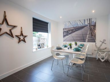 A dining room with a white table set with dishes and glasses and white chairs a window with a black shade star-shaped wall decorations and a large bridge picture at Hafod Heli in Rhosneigr