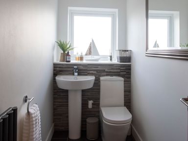 A bathroom with a pedestal sink a toilet a towel rack with a towel a window with decorative items and a mirror on the wall at Hafod Heli in Rhosneigr