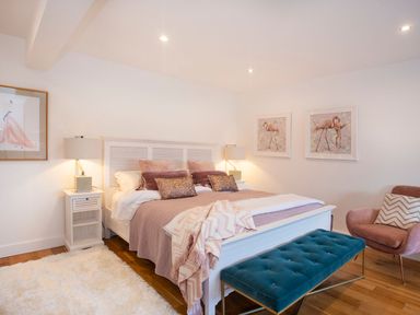 A bedroom with a white bed and pink pillows a teal bench a pink armchair and framed flamingo pictures on the wall at Hafod Heli in Rhosneigr