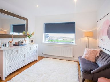 A room with a large white dresser topped with a mirror and decor a gray couch with a pink pillow a floor lamp window with a blue blind and a white rug on a wooden floor at Hafod Heli in Rhosneigr