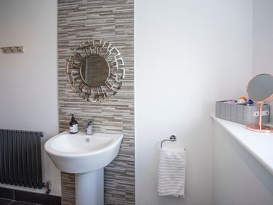 A bathroom with a pedestal sink a round decorative mirror a towel on a rack and toiletries on a windowsill at Hafod Heli in Rhosneigr