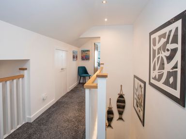A hallway with grey carpet a blue chair wall art and white railing at Hafod Heli in Rhosneigr