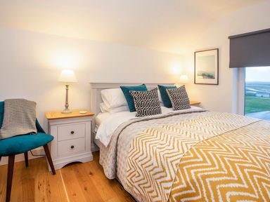A bedroom with a bed with pillows and blankets two bedside tables with lamps a blue chair and a window with a view at Hafod Heli in Rhosneigr