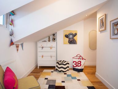 A small room with a white chest of drawers with toys a patterned rug a pouf a bag a yellow chair with a pink pillow and animal artwork at Hafod Heli in Rhosneigr