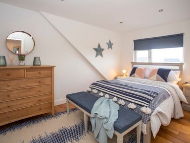 A bedroom with a wooden dresser a bed with blue and white bedding a blue bench and a window with a blind at Hafod Heli in Rhosneigr