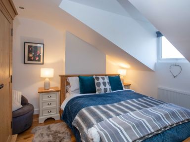 A bedroom with a bed between two bedside tables with lamps a wooden wardrobe a chair with cushions a window and a framed picture on the wall at Hafod Heli in Rhosneigr