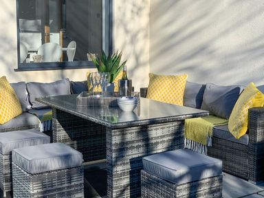 An outdoor seating area with a wicker table and cushioned chairs with yellow pillows at Hafod Heli in Rhosneigr