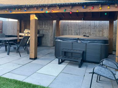 An outdoor covered patio area with a hot tub table grill chairs and string lights at Hafod Heli in Rhosneigr