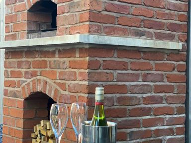 A brick outdoor oven with firewood inside and a wine bottle in a metal ice bucket with two empty wine glasses