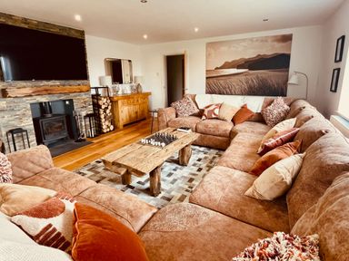 A living room with a large brown sectional sofa and a wooden coffee table with chess pieces a fireplace with a wood-burning stove a flat-screen TV above the fireplace a wooden sideboard with lamps a framed landscape photo on the wall at Hafod Heli in Rhosneigr